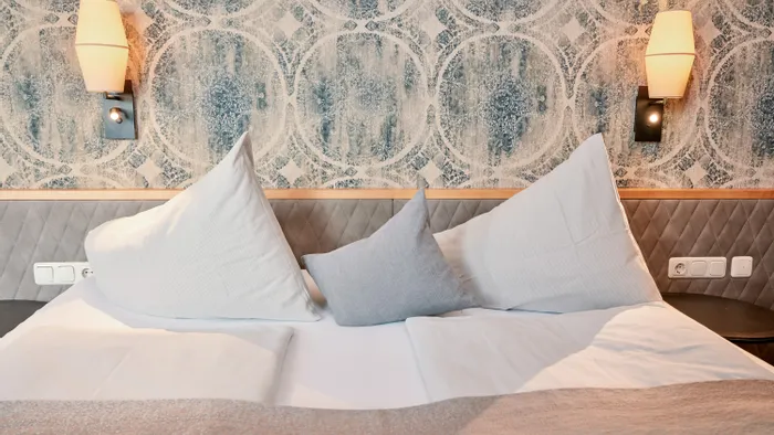 Gemütliches Hotelbett mit großen Kissen, gemusterter Tapete und warmen Wandleuchten